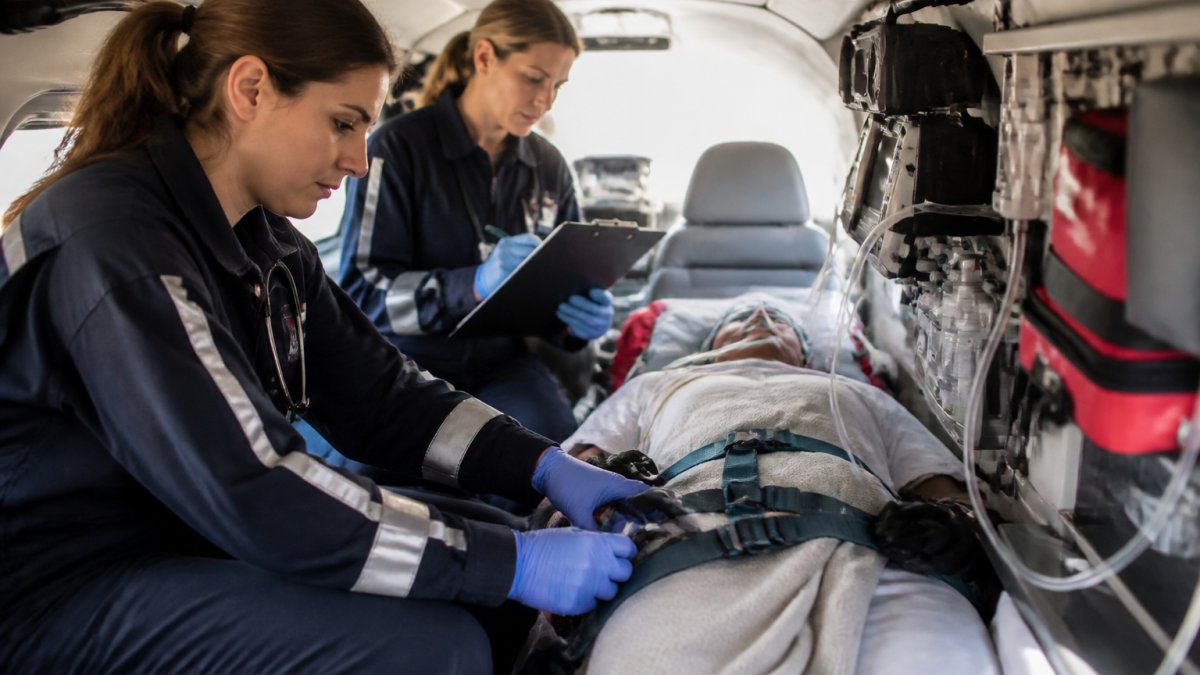 A flight nurse in medical uniform checking monitoring equipment beside a stretcher inside an air ambulance cabin, with a colleague reviewing a clipboard nearby